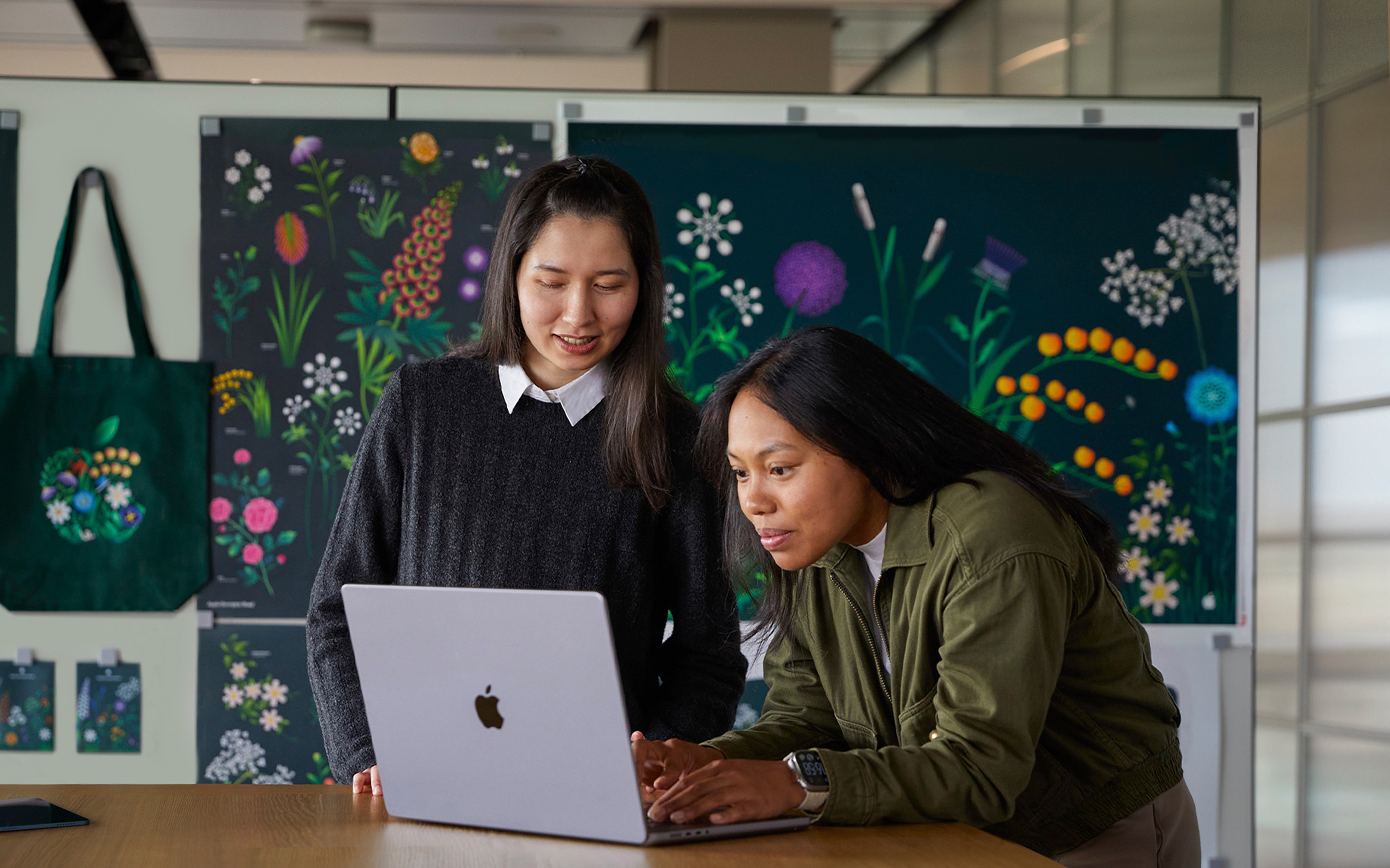 Duas funcionárias da área de marketing trabalhando juntas em um MacBook diante de cartazes criados pela equipe