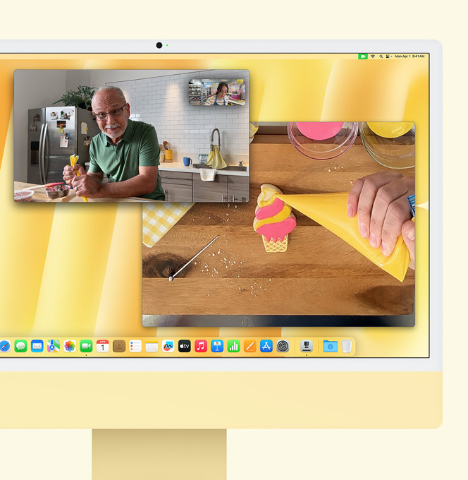 Un iMac giallo con una dimostrazione della funzione Panoramica Scrivania. Sullo schermo c’è una pasticciera in videochiamata con un uomo. Una finestra mostra la pasticciera e il volto dell’uomo. L’altra finestra mostra le mani della pasticciera mentre glassa un biscotto.