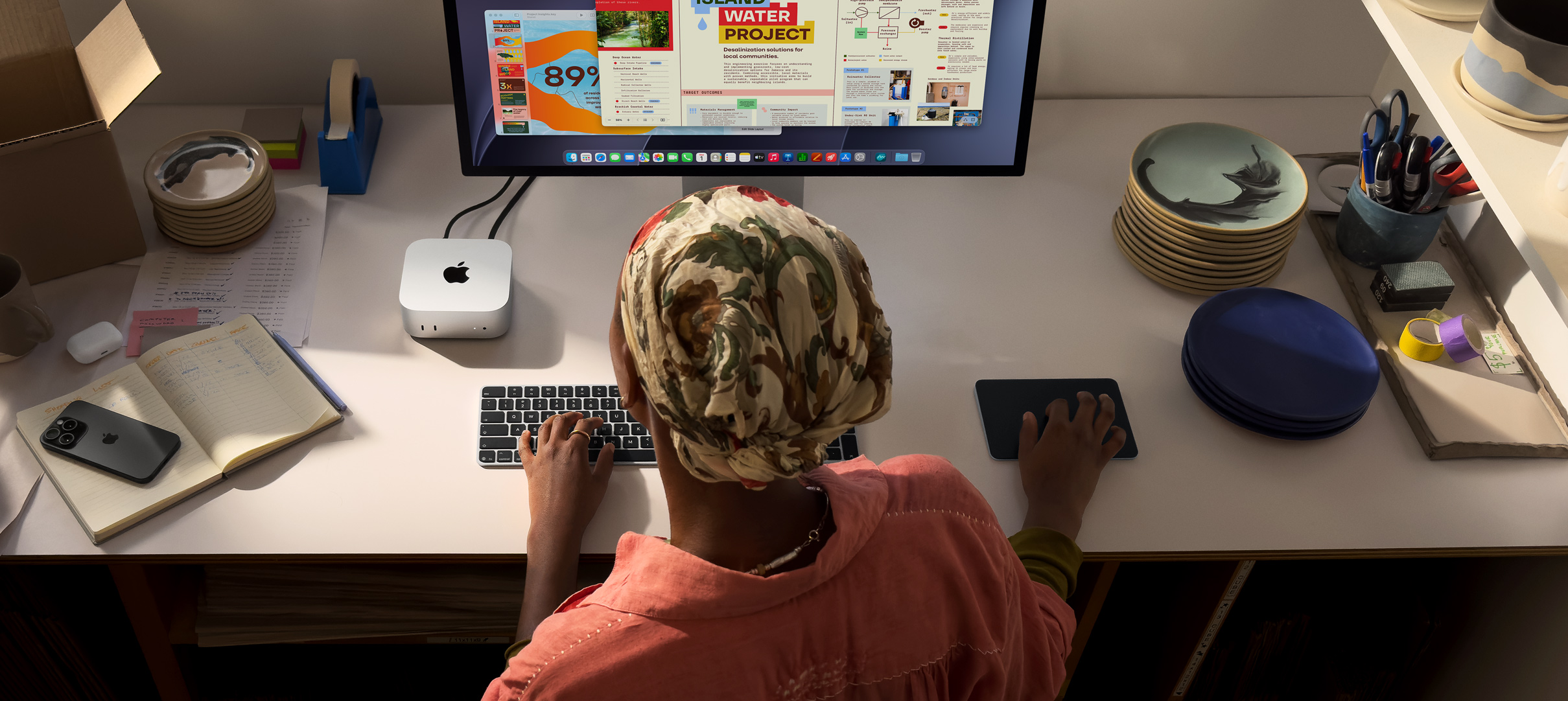 Osoba przy biurku pisze na Macu mini z podłączonym monitorem Studio Display, klawiaturą Magic Keyboard, gładzikiem Magic Trackpad i różnymi przedmiotami