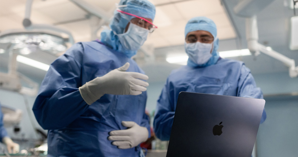 To kirurger på operationsstuen ser på en MacBook, og den ene gestikulerer med hånden, mens den anden ser på