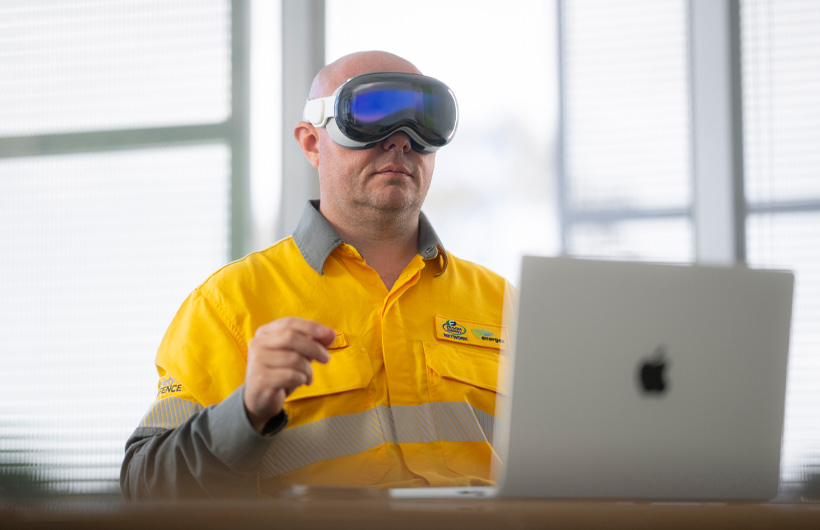 En medarbejder med Apple Vision Pro på laver en knibebevægelse med hånden, mens en sølvfarvet MacBook Pro står åben på skrivebordet