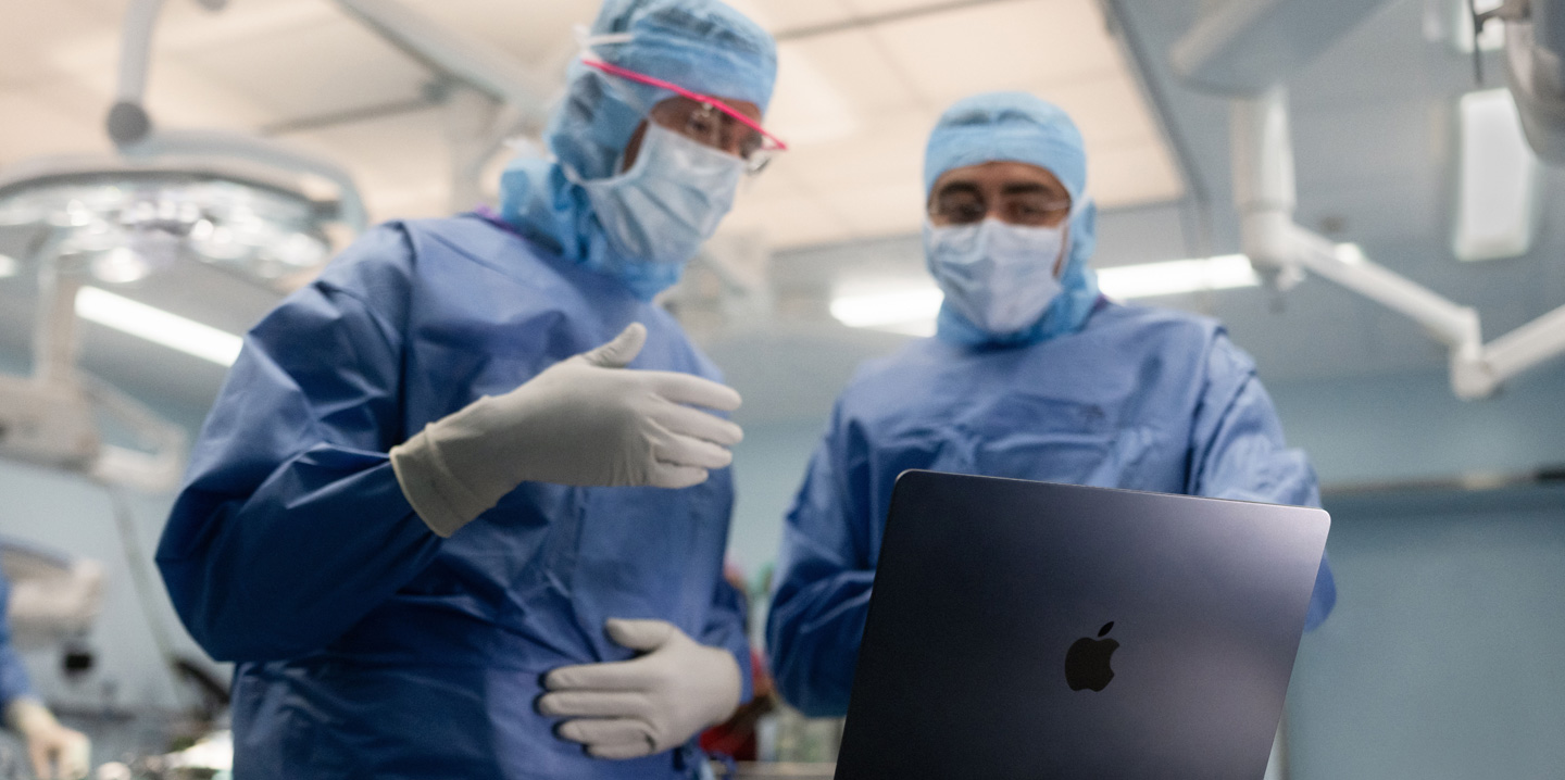 手術室でMacBookを見ている2人の外科医。1人が手でジェスチャーし、もう1人はそれを見ている