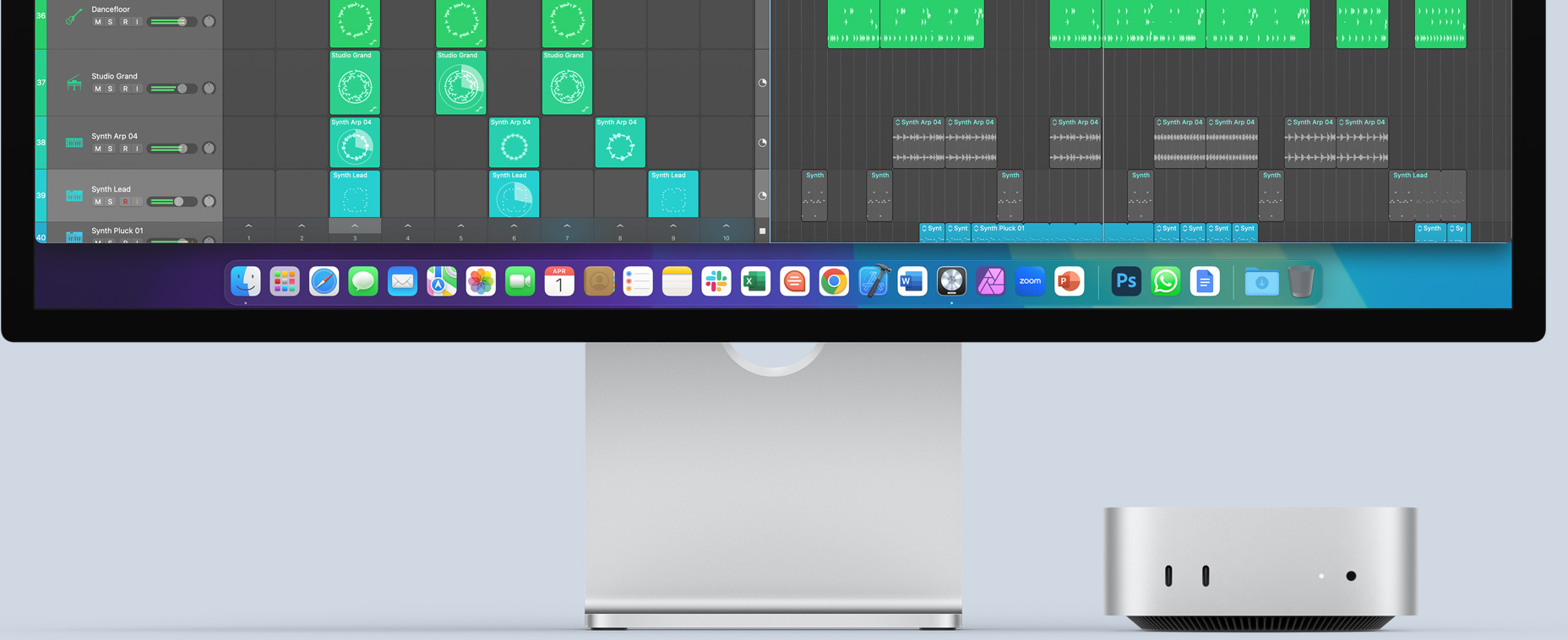 Sidabro spalvos „Mac mini“ priekinė dalis, kurioje matomi priekiniai prievadai ir indikatorinė lemputė, jis įkomponuotas po „Mac“ monitoriumi, kuriame matomas spalvingas ekranas ir doke esančių programų piktogramos